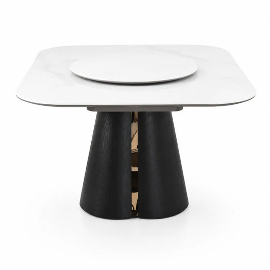 A modern square dining table with a white marble-look top, a round inlay in the center, and a sculptural black pedestal base with a metallic gold accent in the middle, shown on a white background.