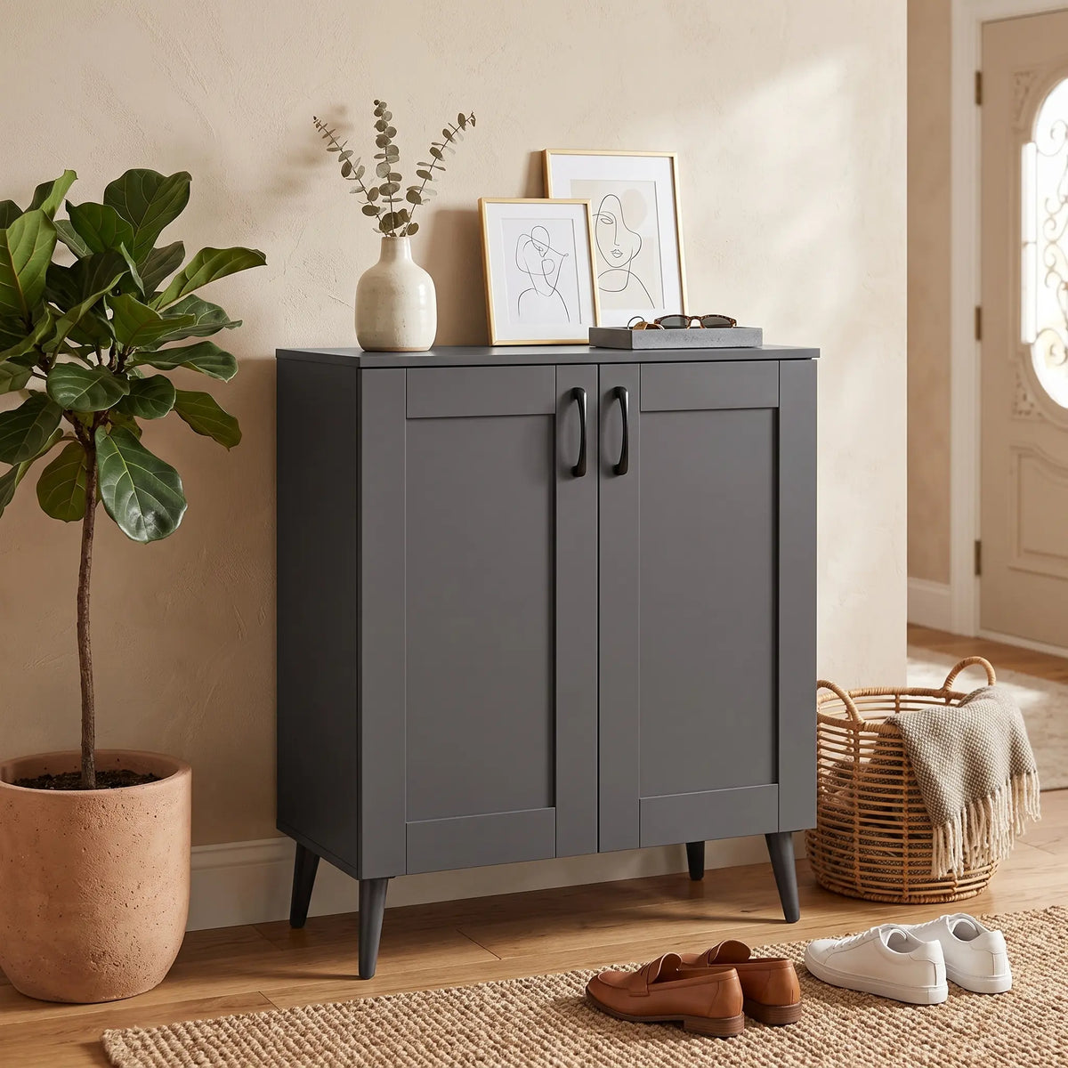 A dark gray two-door cabinet with black handles and tapered legs, styled in a warm, neutral-toned entryway. The cabinet is topped with a small white vase of dried stems and two framed minimalist line drawings.