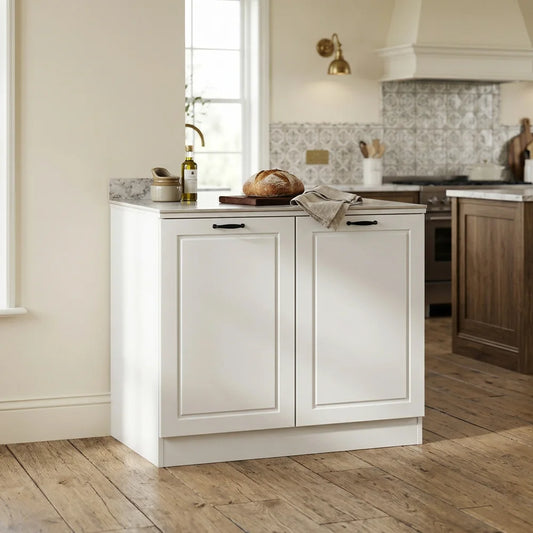 A white kitchen island cabinet with two paneled doors and a light stone countertop, styled with a loaf of bread, a bottle, and a cloth in a bright kitchen setting.