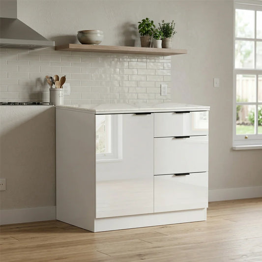 A modern white kitchen cabinet with three drawers and two doors, set in a bright kitchen with a white subway tile backsplash, light wood flooring, and a floating shelf above.