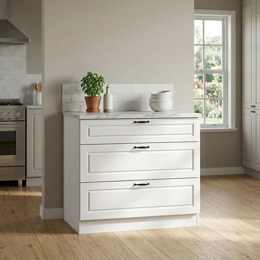 A white three-drawer kitchen cabinet with a light marble-look top and black drawer pulls, shown in a bright kitchen setting with a window, wood floor, and small countertop decor.