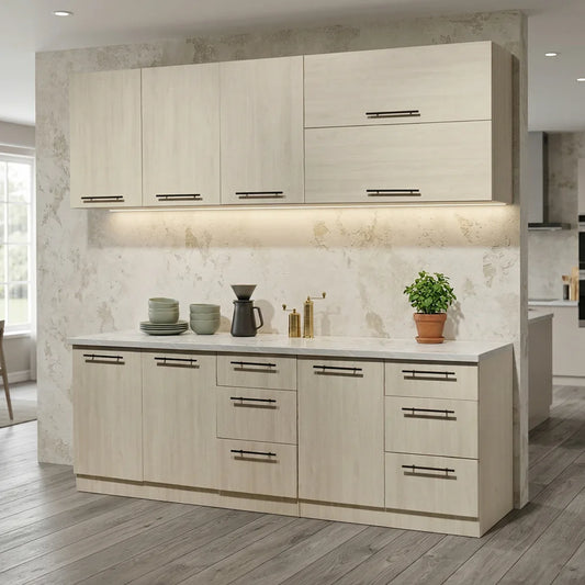 A modern kitchen cabinet and counter set in a light beige wood finish with dark metal handles, set against a textured light gray stone backsplash and wall. The countertop is styled with dishes, a coffee maker, and a small potted plant
