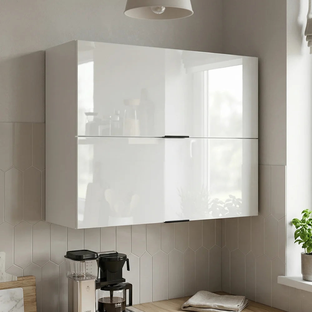 A modern two-door wall-mounted kitchen cabinet in glossy white, installed on a tiled wall above a countertop with a coffee maker and a small plant.